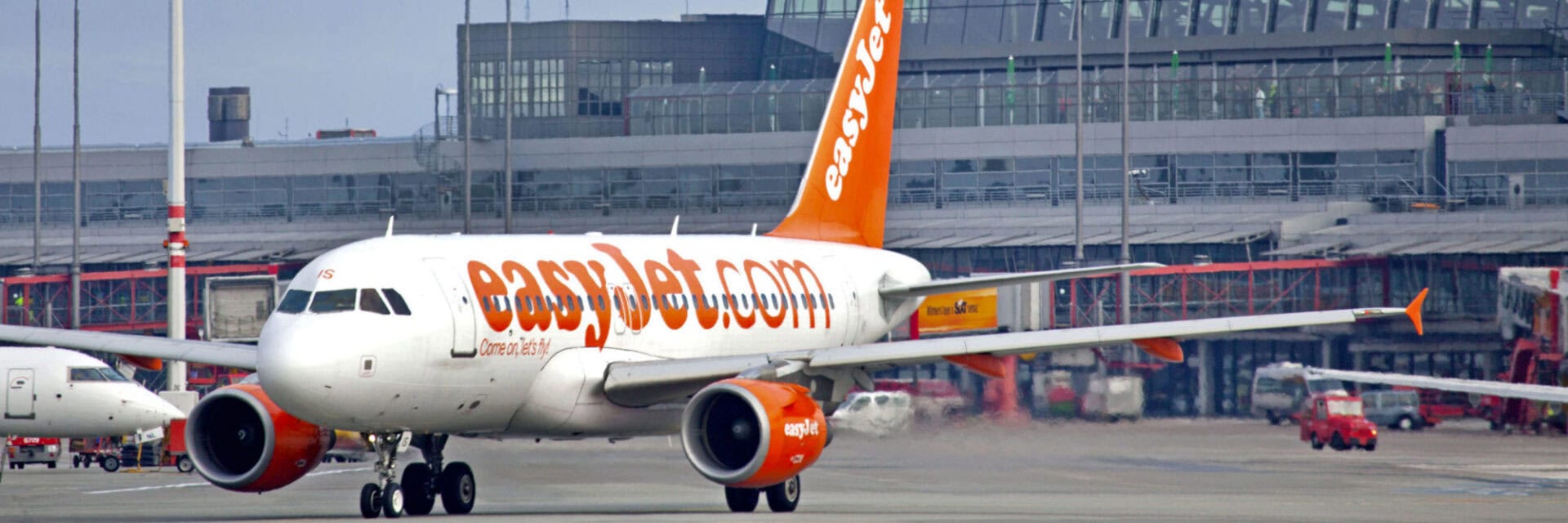 251212-easyJet-hamburg-winterflugplan Ein Flugzeug der Airline Easyjet rollt auf dem Rollfeld eines Flughafens, im Hintergrund ist das moderne Terminal mit Glasfassade zu sehen. Die orange-weiße Lackierung und das große Easyjet-Logo sind gut erkennbar.