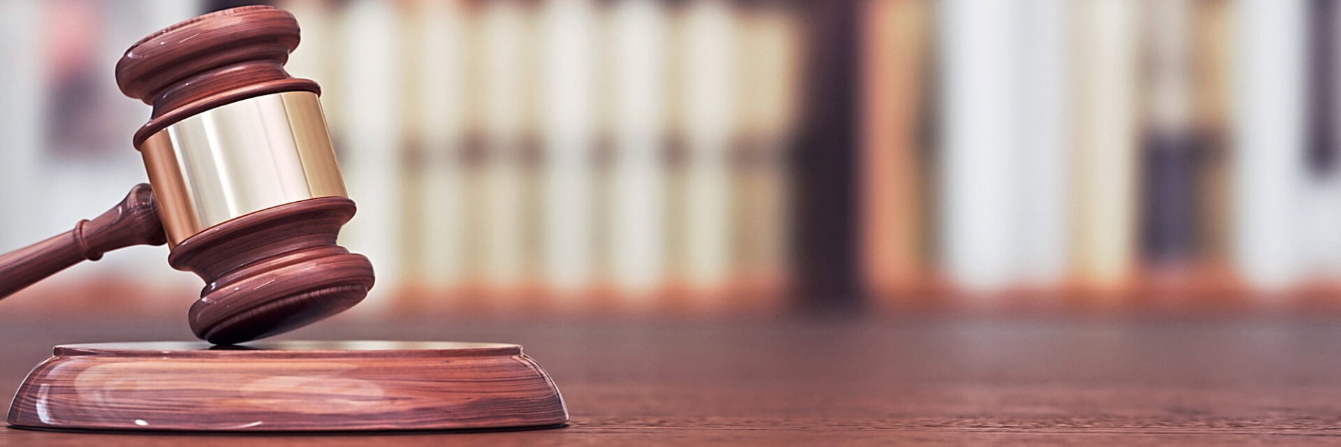 Ein Richterhammer aus Holz liegt auf einem hölzernen Tisch, dahinter ein unscharfes Regal voller Bücher in einer Bibliothek oder einem Gerichtssaal.