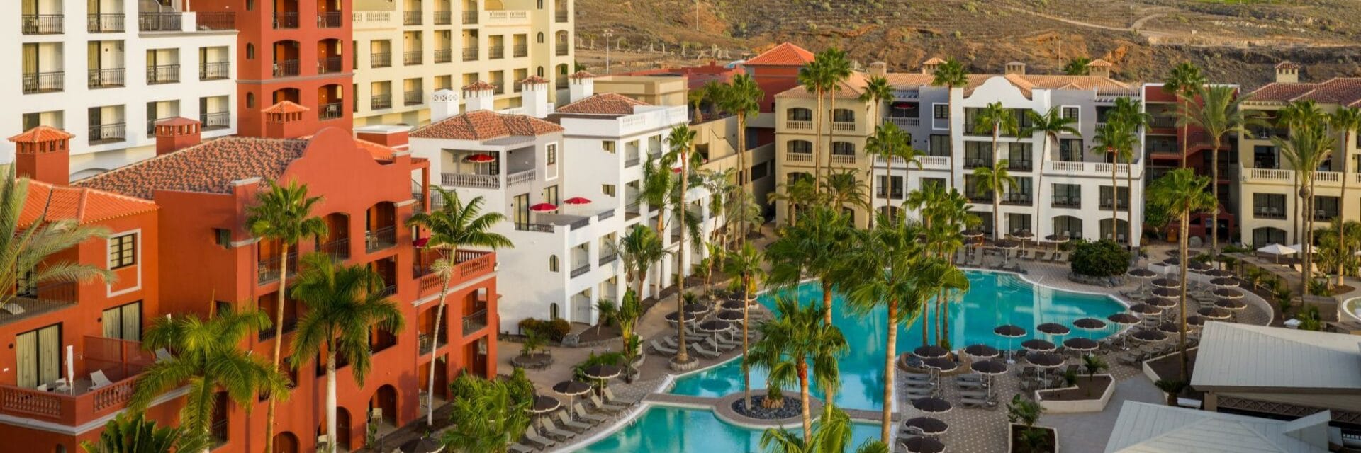 Luftaufnahme eines farbenfrohen Hotelresorts auf Teneriffa mit mehreren Pools, Palmen und umliegenden Bergen – ein ideales Reiseziel für einen entspannten Urlaub auf den Kanaren. Die Gebäude im spanischen Stil umrahmen die große Poollandschaft und bieten zahlreiche Sonnenliegen. Keywords: Hotelresort, Teneriffa, Pool, Palmen, Kanaren, Urlaub, Luftaufnahme.