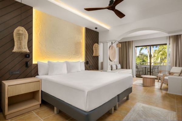 Modernes Hotelzimmer im Resort mit großem Doppelbett, stilvollen Naturmaterialien und Blick auf einen sonnigen Balkon mit Palmen – ideal für Ihren Erholungsurlaub. Die Einrichtung kombiniert helles Holz, Designerlampen und eine gemütliche Sitzecke, die zum Entspannen einlädt