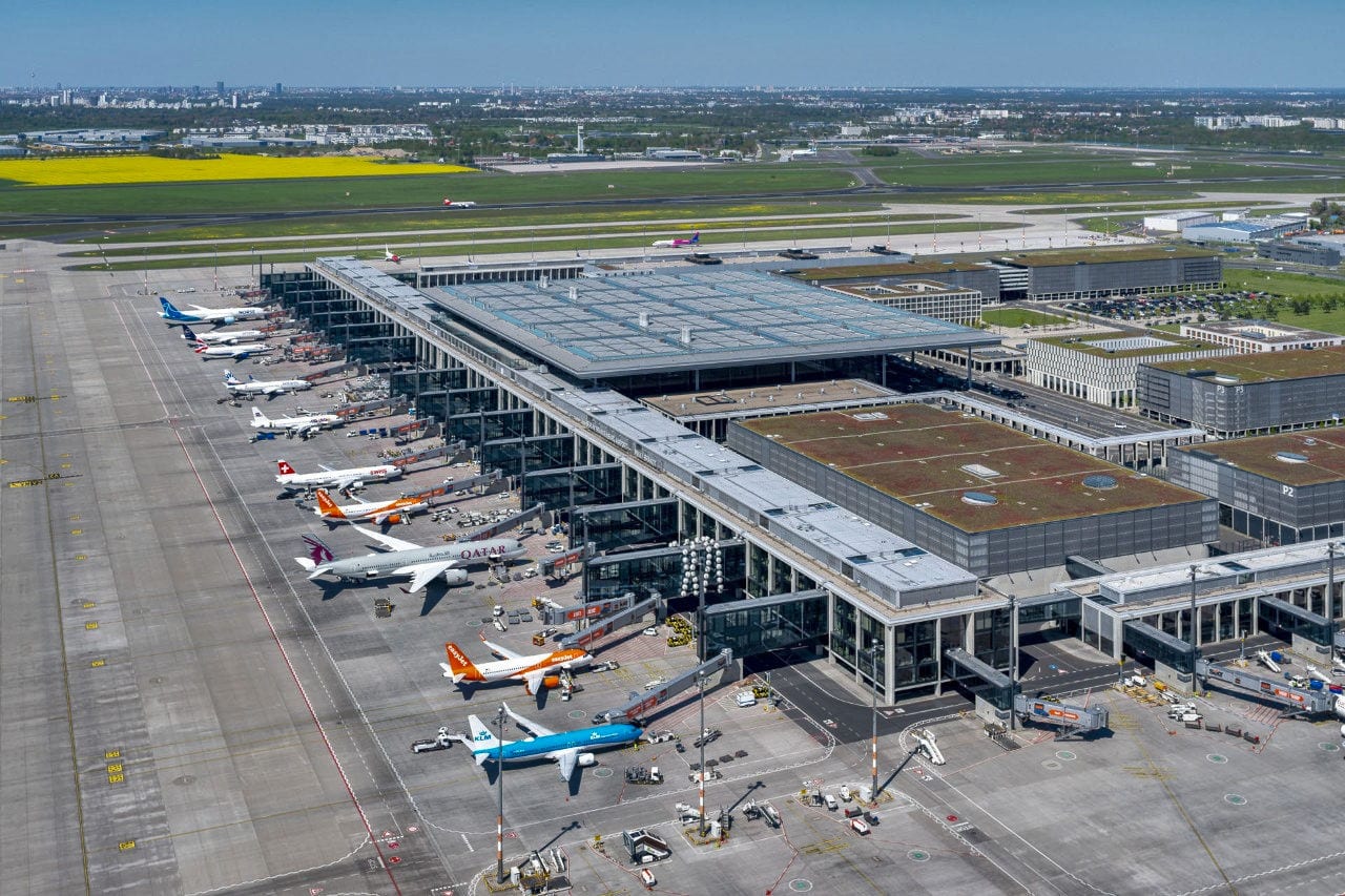Luftaufnahme vom Flughafen BER Berlin Brandenburg mit zahlreichen Flugzeugen verschiedener internationaler Airlines an den Gates, modernen Terminalgebäuden und Start- sowie Landebahnen im Hintergrund.