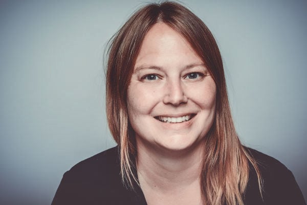 Porträt einer lächelnden Frau mit schulterlangen, hellbraunen Haaren vor einem hellen, neutralen Hintergrund. Sie blickt direkt in die Kamera und trägt ein dunkles Oberteil.