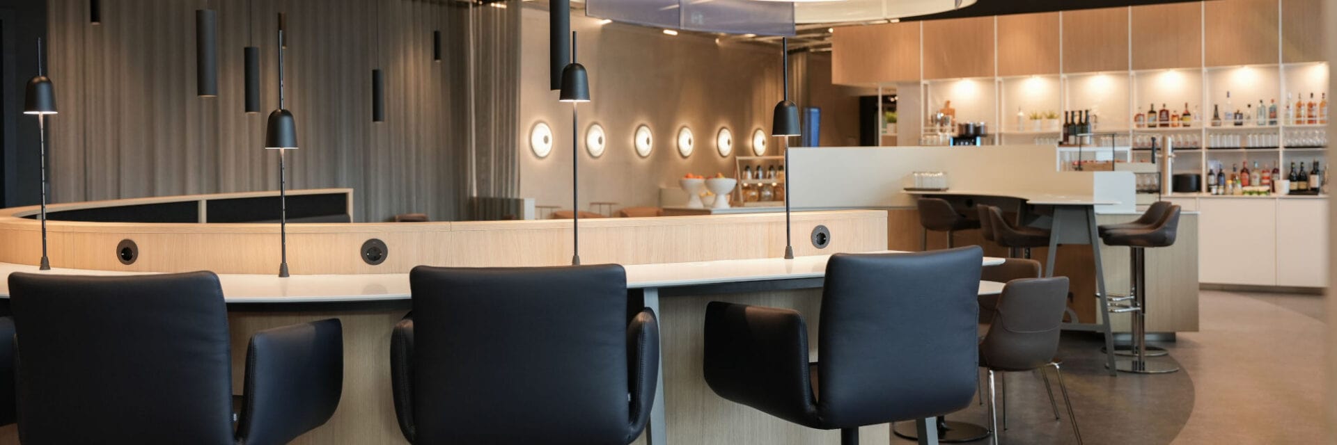 Modern eingerichtete Flughafen-Lounge mit eleganten schwarzen Sesseln, minimalistischen Lampen und einer offenen Bar im Hintergrund, die eine große Auswahl an Getränken präsentiert. Die stilvolle Einrichtung und das warme Licht sorgen für eine entspannte Atmosphäre.