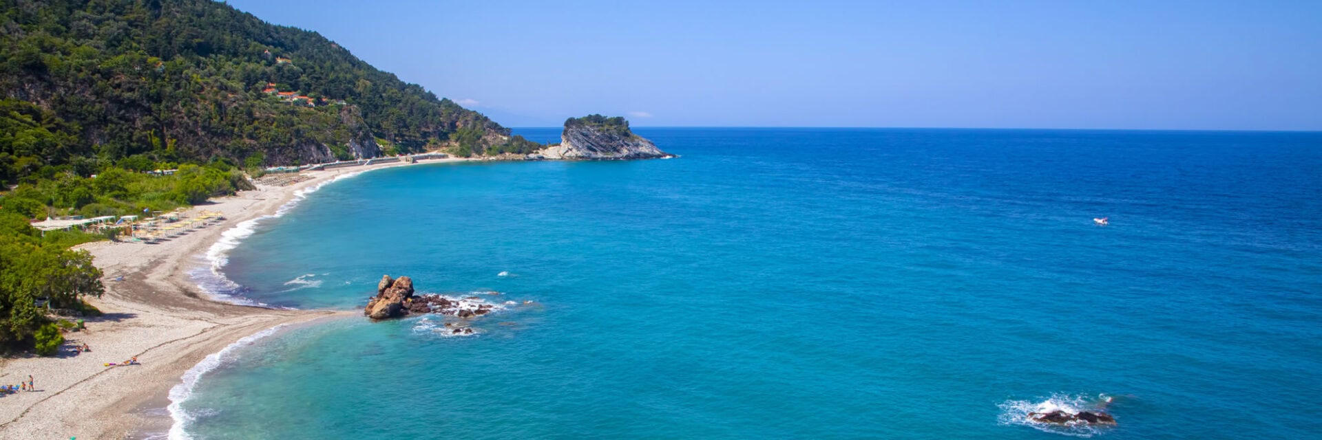 Panoramablick auf eine geschwungene Küste mit hellem Kiesstrand und türkisblauem Meer. Bewaldete Hügel und vereinzelte Felsen rahmen die Bucht ein, im Hintergrund erstreckt sich das offene Meer unter klarem Himmel.