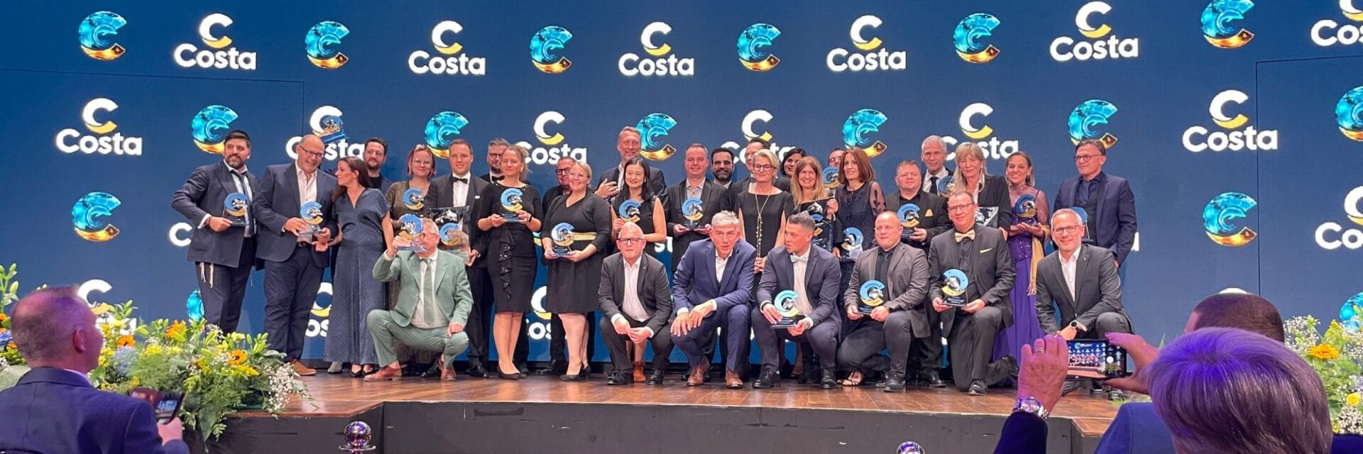 Eine größere Gruppe Menschen steht auf einer Bühne vor einer blauen Wand und hält Preise in der Hand.