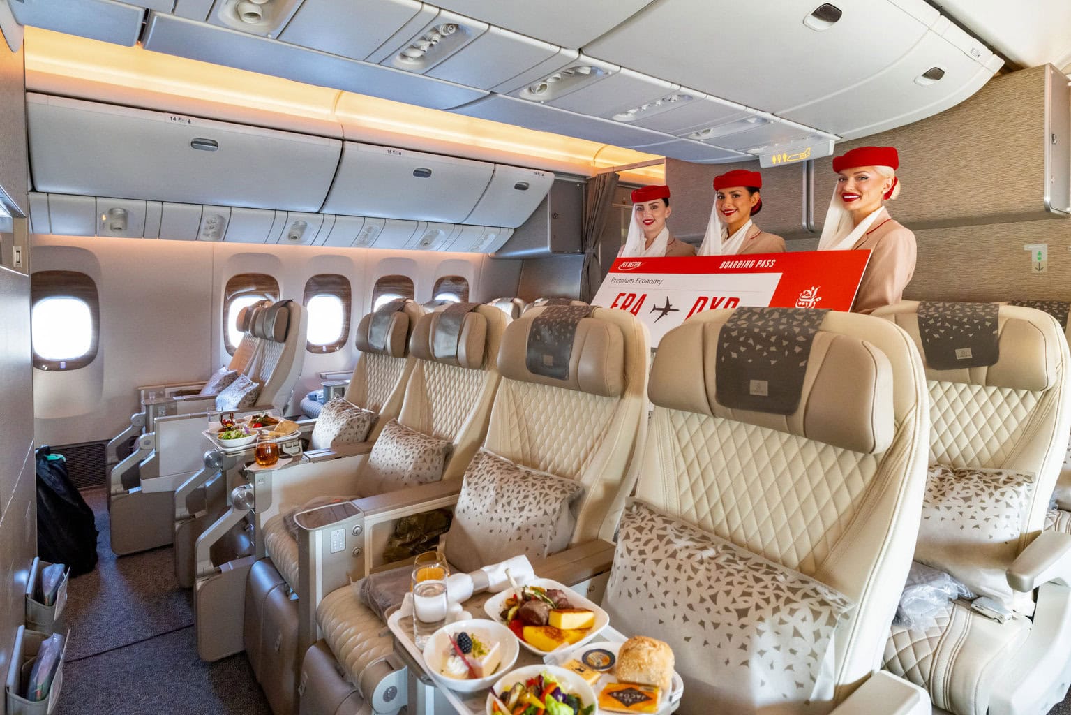 Das Foto zeigt die Premium Economy Kabine eines modernen Passagierflugzeugs mit luxuriösen, hellen Sitzen und gedeckten Tabletts mit Gourmet-Essen. Im Hintergrund stehen drei Flugbegleiterinnen in Uniform, die ein großes Boarding-Pass-Schild präsentieren.