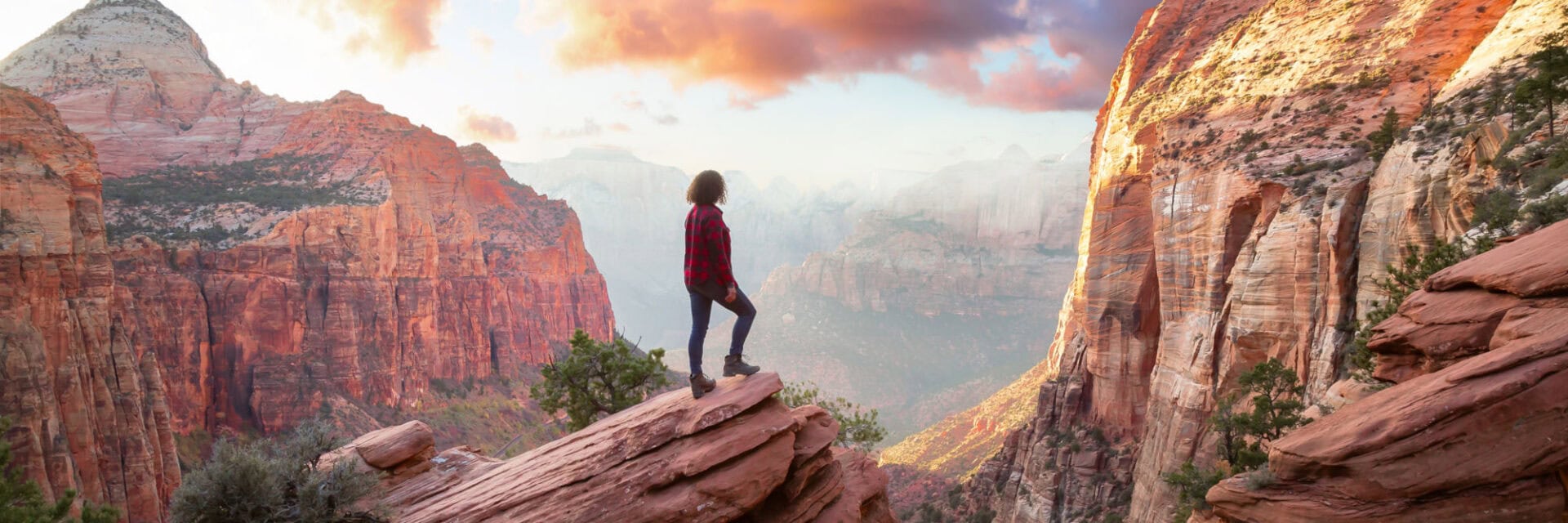 Eine Person steht auf einem Felsvorsprung und blickt in eine weite Schlucht mit hohen, rot leuchtenden Sandsteinfelsen. Die Landschaft ist von Canyons geprägt, darüber ziehen rosafarbene Wolken durch einen hellen Himmel im warmen Licht von Sonnenauf- oder -untergang.