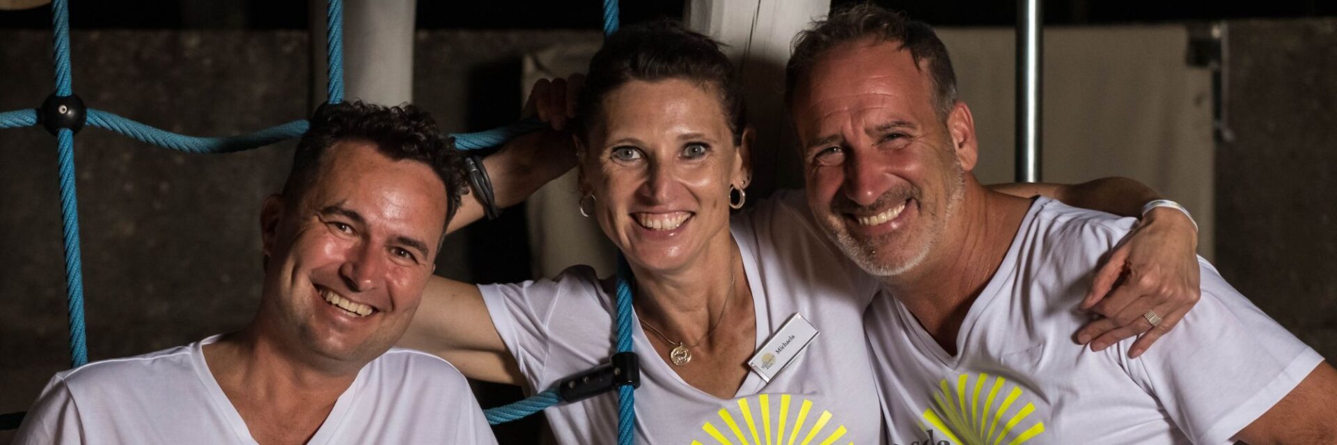 Drei fröhliche Personen stehen nebeneinander und lächeln in die Kamera. Alle tragen weiße T-Shirts mit der Aufschrift „Sola Summer Jam“ und einem gelben Sonnenmotiv. Zwei Männer stehen links und rechts, eine Frau in der Mitte; sie haben jeweils einen Arm umeinander gelegt. Im Hintergrund sind Teile eines Kletternetzes und eine dunkle Wand zu erkennen.