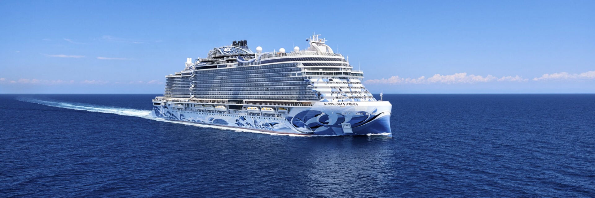Das Foto zeigt ein großes Kreuzfahrtschiff der Norwegian Cruise Line, die Norwegian Prima, das bei strahlend blauem Himmel über das offene Meer fährt. Im Vordergrund ist das klare Wasser zu sehen, während das moderne Schiff mit auffälligem Design Richtung Horizont steuert.
