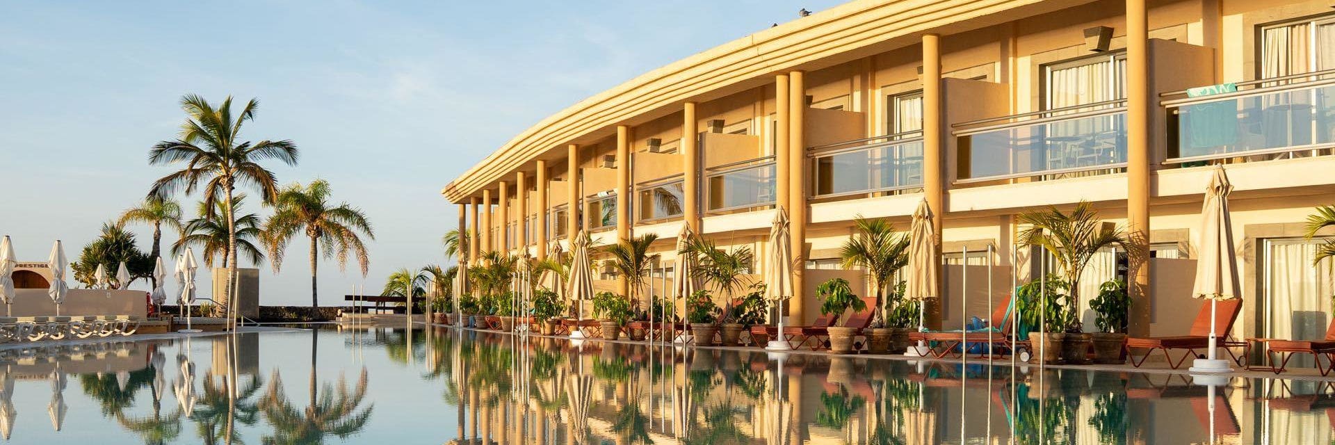 251218-schauinsland-inforeisen Modernes Hotelresort mit geschwungener Fassade und Balkonen, das sich in einem ruhigen, spiegelglatten Pool widerspiegelt; Palmen, Sonnenschirme und Liegen am Pool bei warmem Abendlicht unter blauem Himmel.