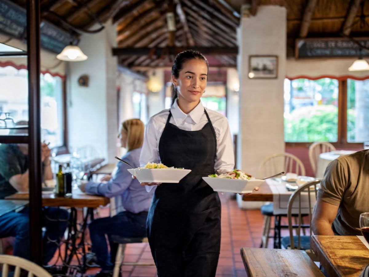 Eine Kellnerin im deutschen Restaurant serviert zwei Gerichte an Gäste, die entspannt an Holztischen sitzen. Die gemütliche Gastronomie mit rustikalem Interieur und großen Fenstern ist gut besucht.
