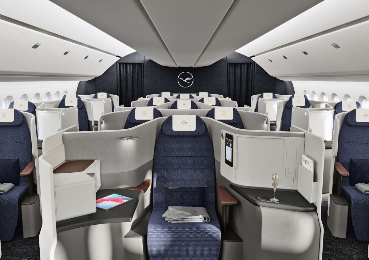 Die Aufnahme zeigt das Interieur der Lufthansa Business Class mit geräumigen Sitzen, modernen Ablagen und einem eleganten Kabinendesign; ein Glas Champagner und ein Tablet unterstreichen das luxuriöse Ambiente. Die Sitzreihen bieten viel Platz und Privatsphäre in einer hellen, freundlichen Flugzeugkabine.