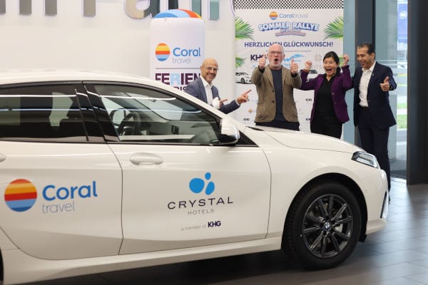 Vier jubelnde Personen stehen in einem Reisebüro neben einem weißen BMW mit den Logos von Coral Travel und Crystal Hotels; im Hintergrund sind Werbebanner zur „Coral Travel Sommer Rallye“ zu sehen.