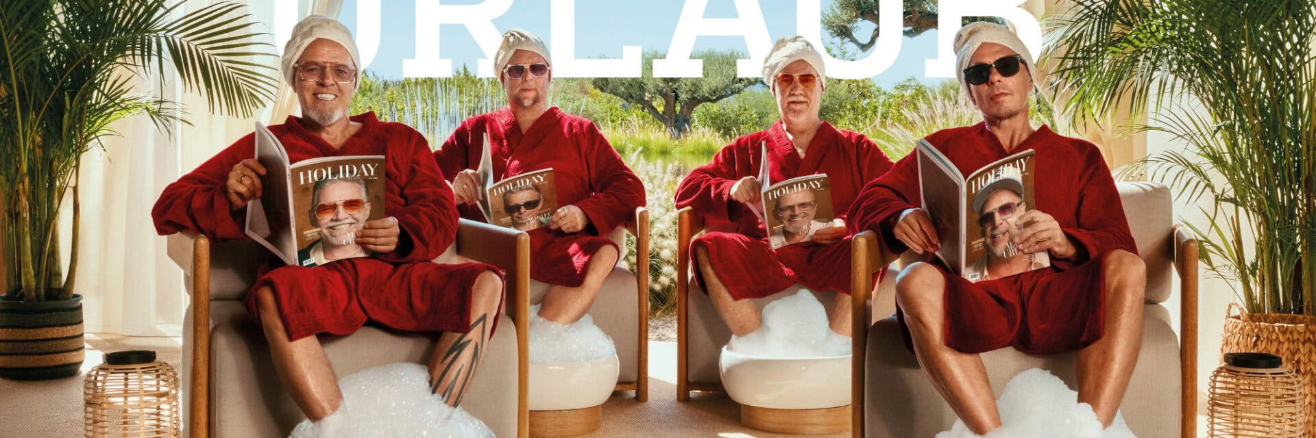 Vier erwachsene Männer in roten Bademänteln sitzen nebeneinander in Sesseln, tragen Handtücher und Sonnenbrillen, lesen Magazine und haben die Füße in schaumgefüllten Fußbädern in einer hellen, grünen Wellness-Umgebung.