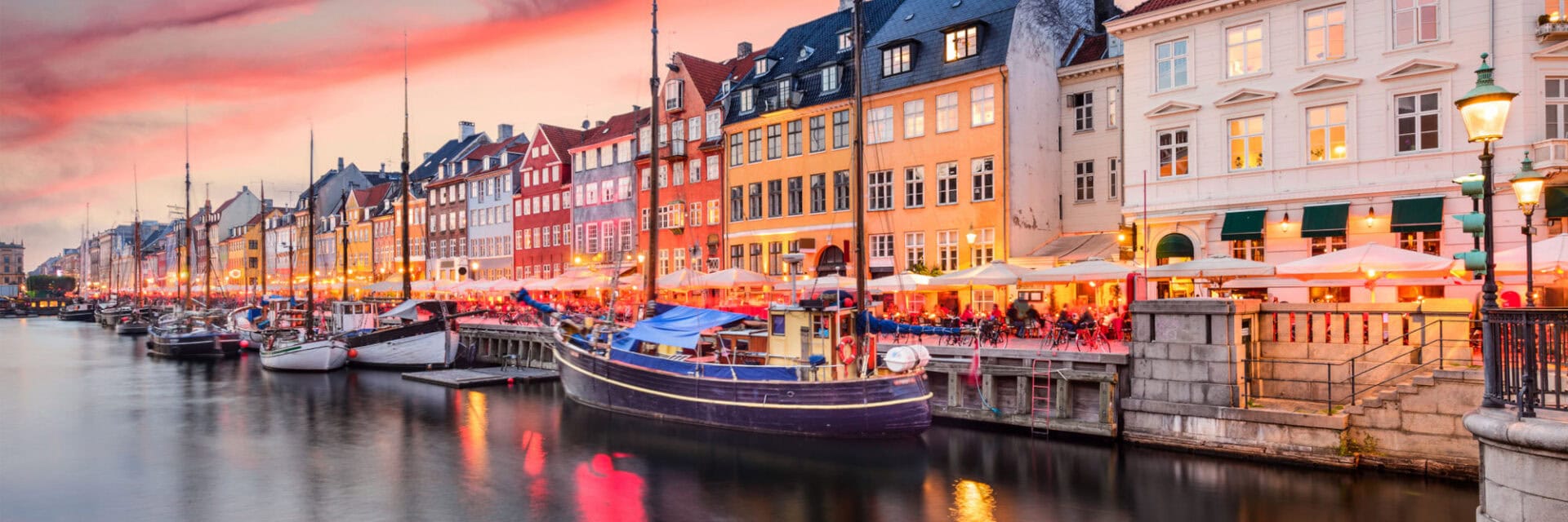 Abendliche Hafenszene mit bunten historischen Häusern entlang eines Kanals; beleuchtete Straßencafés am Ufer, mehrere Boote im Wasser und warme Lichter, die sich bei Sonnenuntergang im Kanal spiegeln.