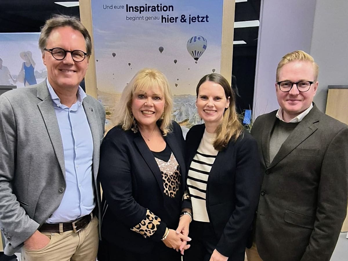Vier Personen stehen lächelnd nebeneinander in einem Büro vor einem Lufthansa City Center Banner mit der Aufschrift „Und eure Inspiration beginnt genau hier & jetzt“; zwei Männer im Anzug links und rechts sowie zwei Frauen im Business Outfit in der Mitte, im Hintergrund eine Landschaft mit Heißluftballons.
