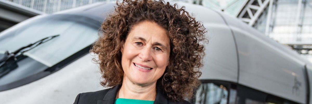 Eine Managerin mittleren Alters mit dunklem Sakko und grüner Bluse steht lächelnd vor einem ICE-Zug vor einem Bahnhofsgebäude.