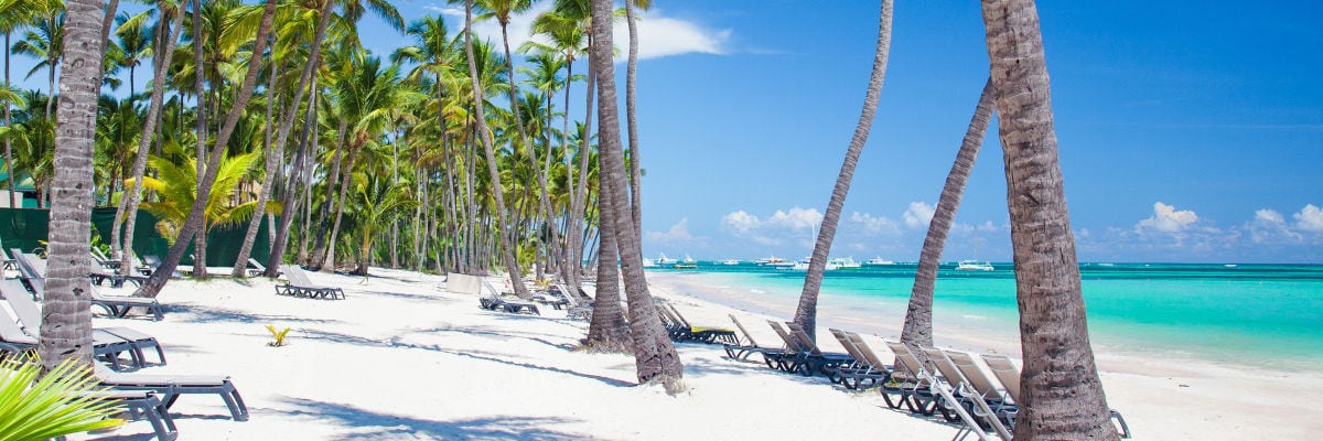 Liegestuhl am Strand mit Blick auf türkisfarbenes Meer, Palmen wiegen sich im Wind.