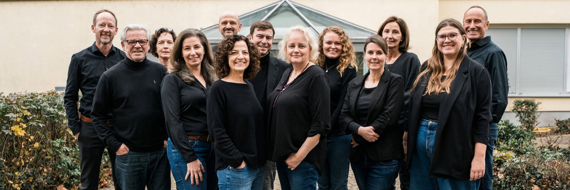 Eine Gruppe von dreizehn Personen steht dicht beisammen im Freien vor einem Gebäude mit einem verglasten Dachaufsatz. Alle tragen dunkle Oberteile und überwiegend Jeans. Die Gruppe lächelt in die Kamera, und im Vordergrund sind Sträucher und herbstliche Bepflanzung zu sehen.