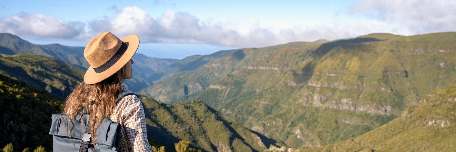 Das Foto zeigt eine Frau mit einem Wanderrucksack auf dem Rücken von hinten, die über eine grüne Hügellandschaft blickt.