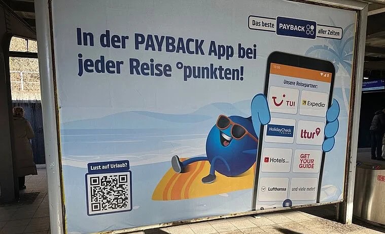 Werbeplakat am Bahnhof für die PAYBACK App: Links groß der Text „In der PAYBACK App bei jeder Reise °punkten!“, darunter ein QR-Code mit der Frage „Lust auf Urlaub?“; rechts sitzt das blaue PAYBACK-Maskottchen mit Sonnenbrille auf einem Surfbrett und hält ein großes Smartphone hoch, auf dessen Display verschiedene Reisepartner wie TUI, Expedia, HolidayCheck, ltur, Hotels.com, Lufthansa und GetYourGuide angezeigt werden.