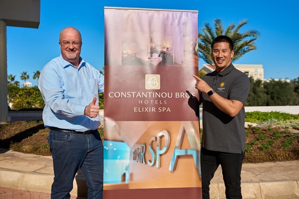 Zwei Männer stehen neben einem Aufsteller der Constantinou Bros Hotels und halten den Daumen in die Kamera