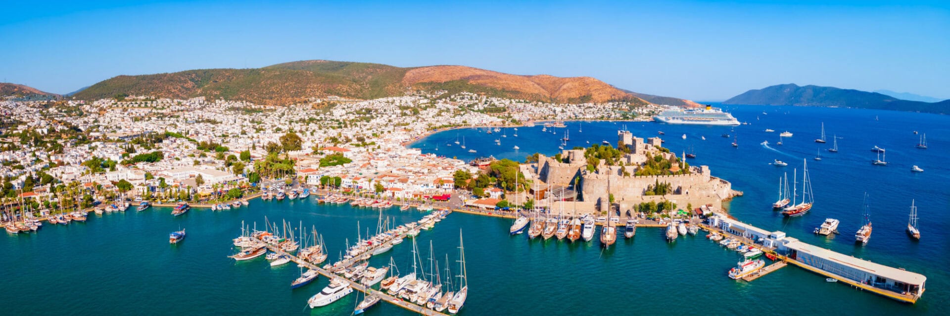 Panoramablick auf eine Küstenstadt mit weiß getünchten Häusern, einem historischen Steinschloss am Hafen und zahlreichen Segelbooten und Yachten im tiefblauen Meer; dahinter hügelige Landschaft unter klarem, blauem Himmel