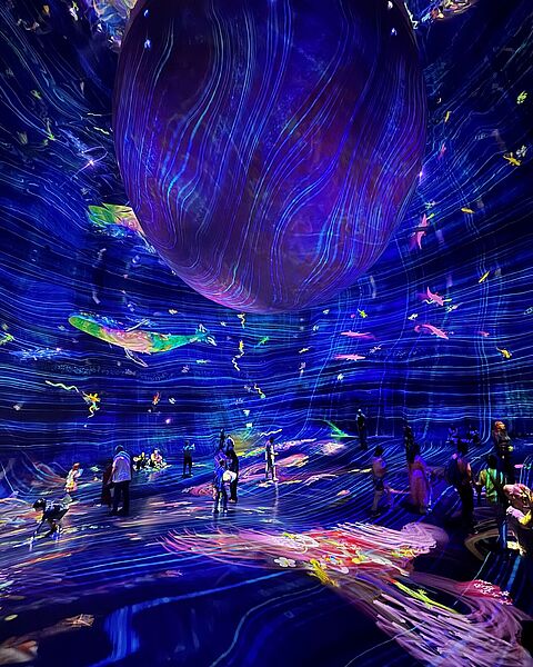 In einer futuristisch gestalteten Halle mit leuchtenden, blauen Lichtern und abstrakten, farbenfrohen Projektionen bewegen sich zahlreiche Menschen durch eine immersive Kunstinstallation; der Fokus liegt auf einer riesigen, schwebenden Kugel in der Mitte