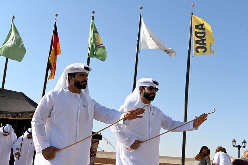 Zwei Männer in traditioneller arabischer Kleidung tanzen mit Stöcken unter strahlend blauem Himmel, im Hintergrund wehen mehrere Fahnen, darunter die deutsche Flagge und eine gelbe ADAC-Fahne