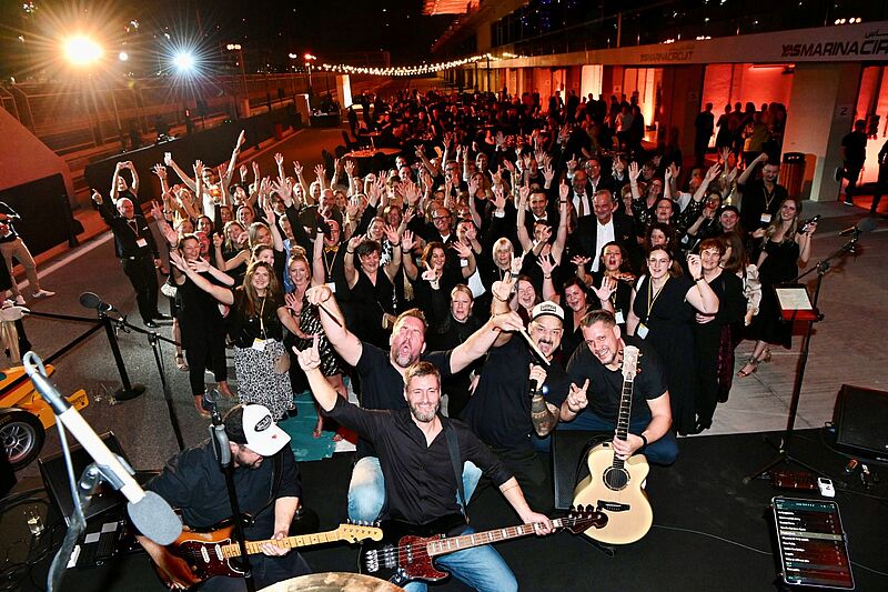 Große Menschenmenge bei einer ausgelassenen Abendveranstaltung im Freien; im Vordergrund posiert eine Liveband mit Gitarren und Mikrofonen, während die feiernden Gäste hinter ihnen fröhlich die Hände in die Luft werfen