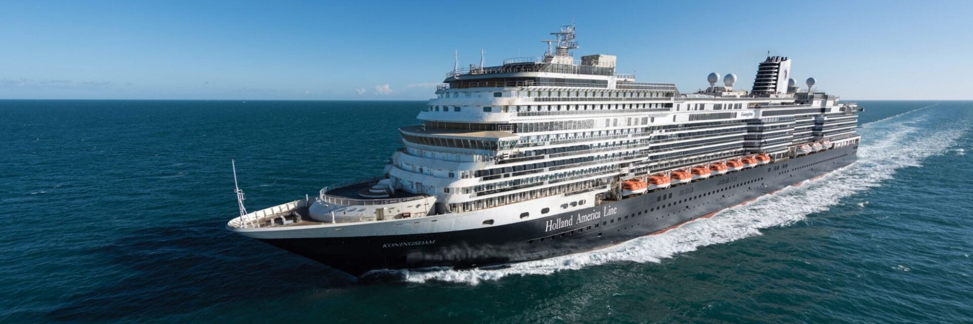 Großes Kreuzfahrtschiff der Holland America Line fährt bei klarem blauem Himmel über das offene Meer; das Schiff erzeugt eine weiße Bugwelle, während es von links nach rechts durchs Wasser gleitet.“