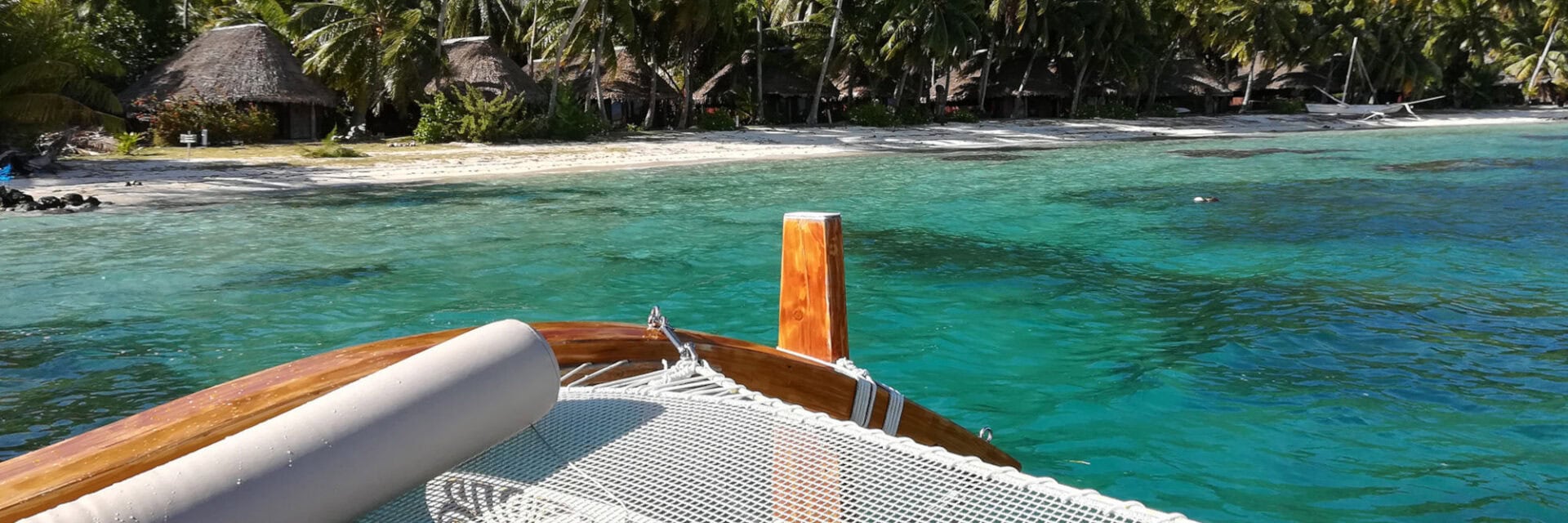 Blick vom Bug eines Bootes auf eine tropische Insel mit weißem Sandstrand, türkisblauem Wasser, Palmen und strohgedeckten Hütten entlang der Küste.