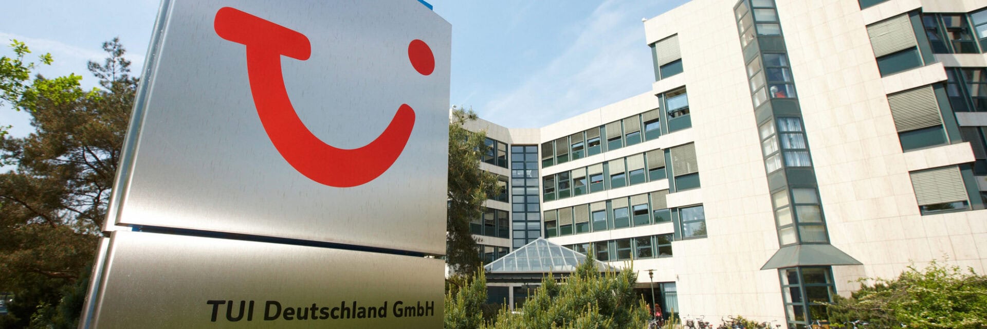Großes Firmenschild mit rotem TUI-Logo und dem Schriftzug „TUI Deutschland GmbH“ vor einem modernen Bürogebäude mit vielen Fenstern und Glasentrée, umgeben von Bäumen.