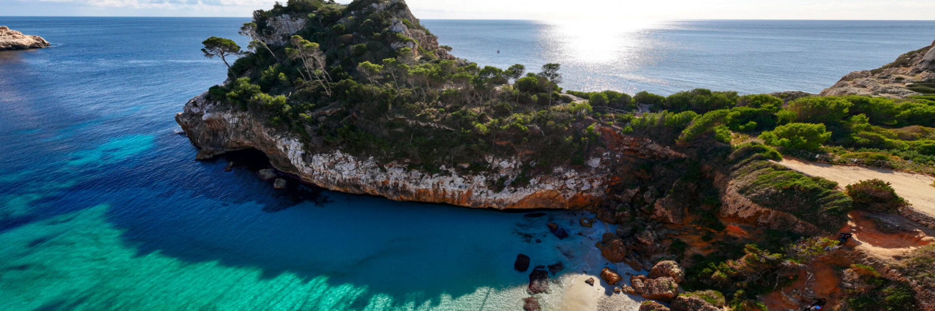 Felsige Mittelmeerküste mit türkisblauem, klarem Wasser, einer kleinen geschützten Bucht und bewachsenen Klippen; oben verläuft ein schmaler Weg entlang der Küste, im Hintergrund offenes Meer und Sonnenlicht auf der Wasseroberfläche.