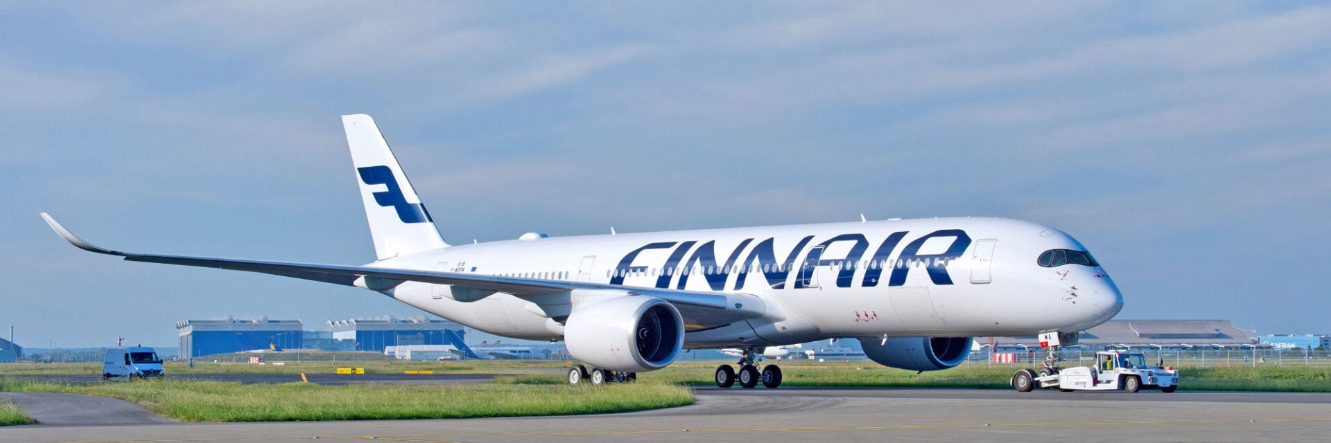 Ein großes Finnair-Flugzeug, ein moderner Airbus auf dem Rollfeld eines Flughafens, wird von einem Pushback-Fahrzeug bewegt. Im Hintergrund sind Flughafenhangars und weitere Flugzeuge zu sehen.