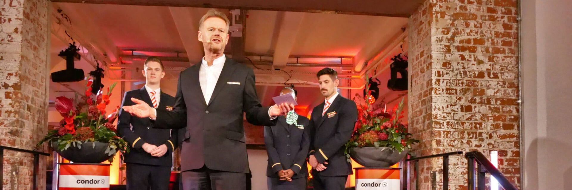 Vier Personen in Uniformen und Anzug stehen auf einer Bühne bei einer Veranstaltung der Fluggesellschaft Condor, umgeben von großen Blumenarrangements und Backsteinwänden; das Bild zeigt eine feierliche Präsentation oder Ansprache.