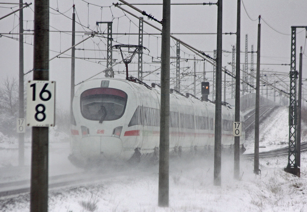 Ein ICE-Schnellzug der Deutschen Bahn fährt bei winterlichem Schneetreiben durch eine verschneite Landschaft, umgeben von Oberleitungen und Signalmasten. Die Bahnstrecke ist mit Schneeverwehungen bedeckt, was die Herausforderungen für den Zugverkehr im Winter zeigt.