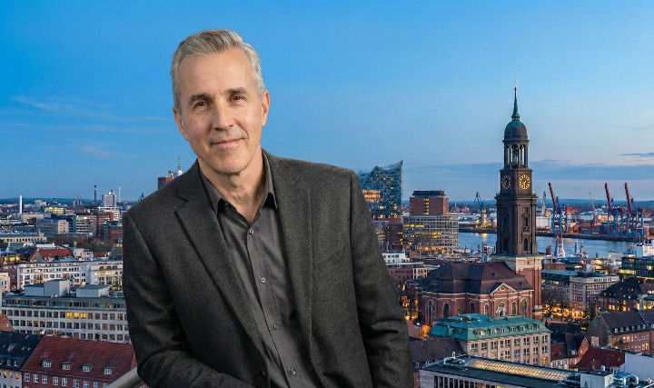Im Vordergrund sitzt ein Mann im dunklen Sakko, während im Hintergrund die berühmten Sehenswürdigkeiten Hamburgs wie der Michel und der Hafen bei Sonnenuntergang zu sehen sind.
