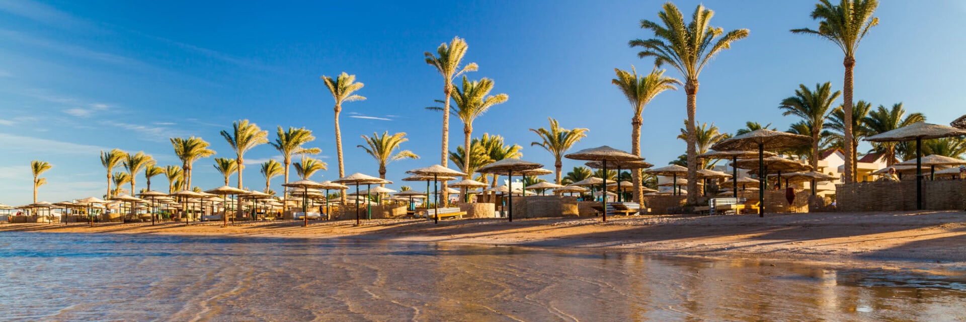 Palmengesäumter Sandstrand mit Reihen von Sonnenschirmen und Liegen an flachem, klarem Wasser, dahinter eine Hotelanlage unter blauem Himmel.