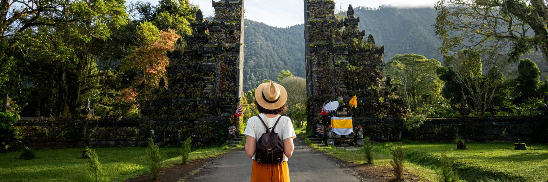 Touristin mit Rucksack steht vor einem balinesischen Tempeltor in grüner Landschaft auf Bali.