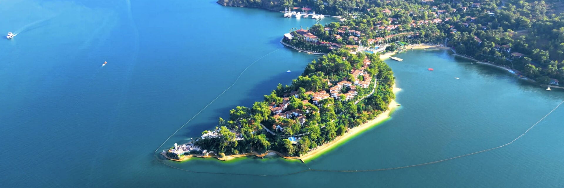 Luftaufnahme einer grünen Halbinsel mit roten Ziegeldächern, umgeben von türkisblauem Meer, kleinen Sandstränden, Booten und einer Marina.