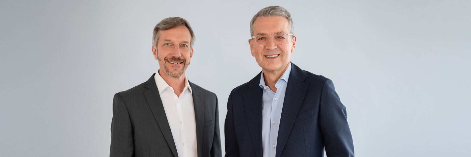 Zwei Männer in Businesskleidung im mittleren Alter stehen nebeneinander vor hellem Hintergrund und lächeln in die Kamera.