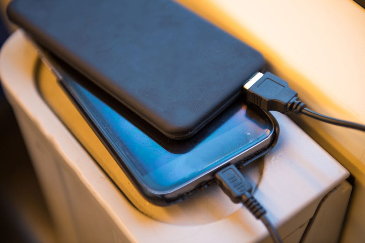 Zwei Smartphones liegen übereinander auf einer schmalen Ablage, vermutlich in einem Flugzeug. Beide Geräte sind über Kabel mit schwarzen Powerbanks verbunden und werden geladen. Das obere Smartphone verdeckt teilweise das untere, dessen Display blau leuchtet.