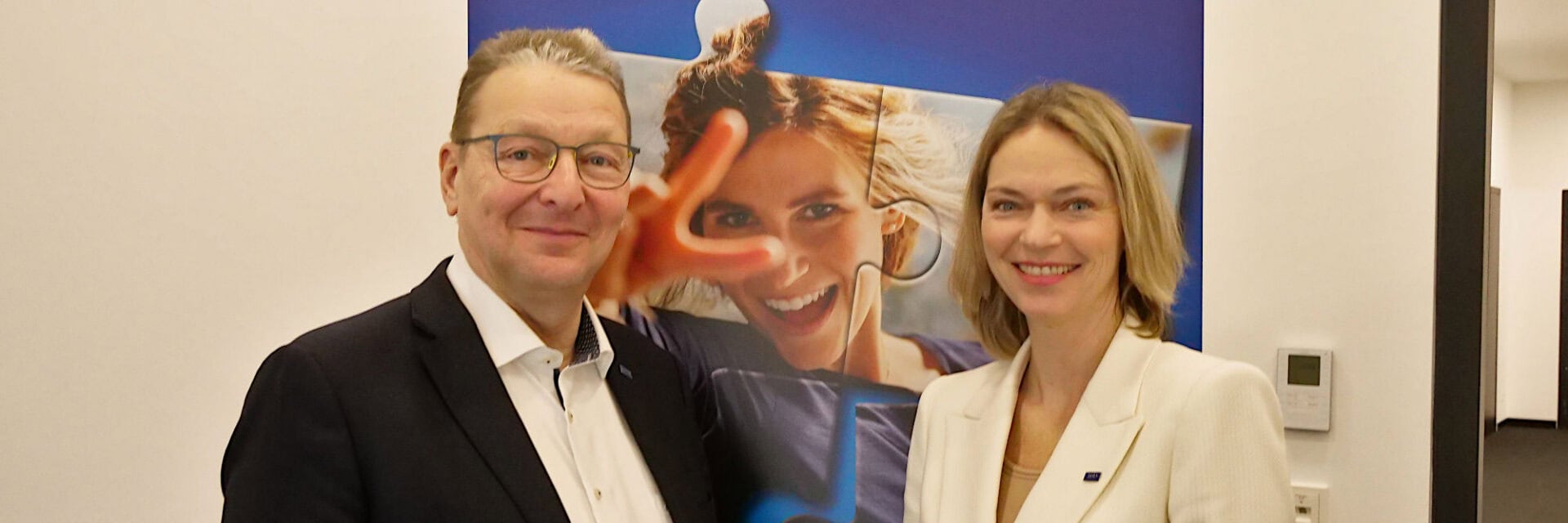 Ein älterer Manager mit dunklem Sakko und weißem Hemd steht neben einer jungen Managerin mit hellem Blazer. Beide stehen lächelnd vor einer Werbewand mit einer lachenden jungen Frau auf einem Puzzlestück.