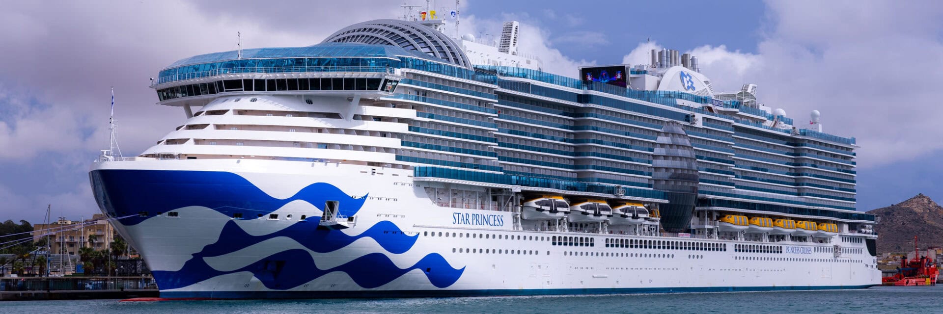 Ein großes Kreuzfahrtschiff mit dem Namen „Star Princess“ liegt am Pier im Hafen von Cartagena, Spanien. Das moderne Kreuzfahrtschiff von Princess Cruises beeindruckt mit mehreren Decks, zahlreichen Balkonkabinen und Rettungsbooten an der Seite. Im Hintergrund sind der Hafen, Gebäude und Hügel unter bewölktem Himmel zu sehen.