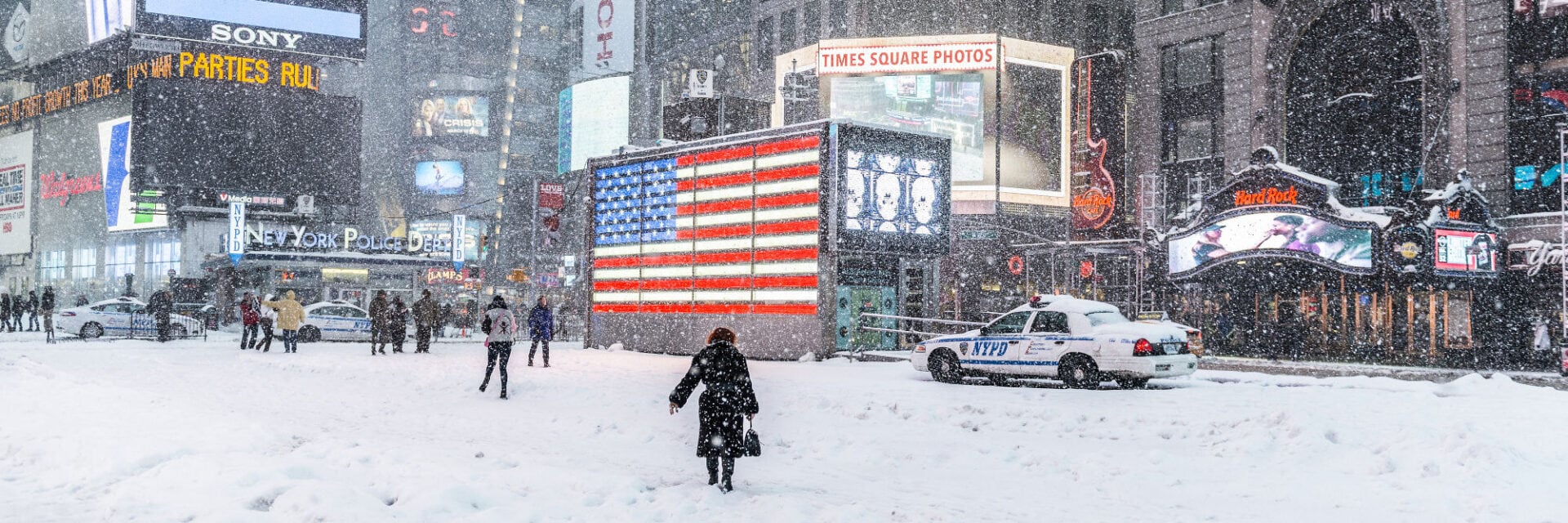 260126-usa-wintersturm-times-square-new-york Das Bild zeigt den berühmten Times Square in New York City während eines heftigen Wintersturms mit dichtem Schneefall. Menschen laufen durch die verschneiten Straßen, vor einer leuchtenden amerikanischen Flagge und einem NYPD-Polizeiauto.