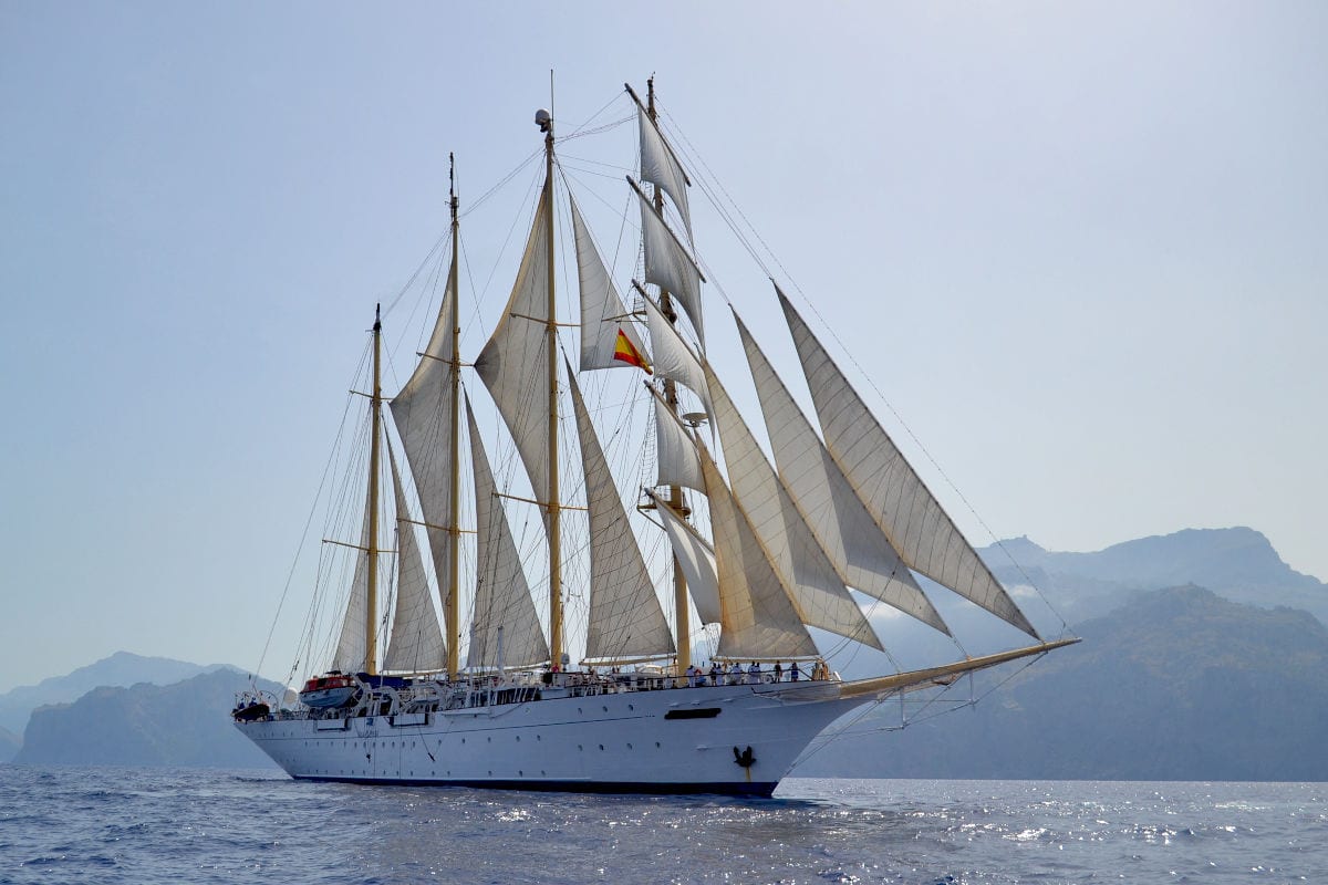 Ein großes, weißes Segelschiff mit mehreren Masten und vollen Segeln fährt majestätisch auf dem offenen Meer, im Hintergrund sind Berge zu sehen. Das Bild zeigt eine klassische Segelyacht auf Kreuzfahrt vor einer mediterranen Küstenlandschaft.