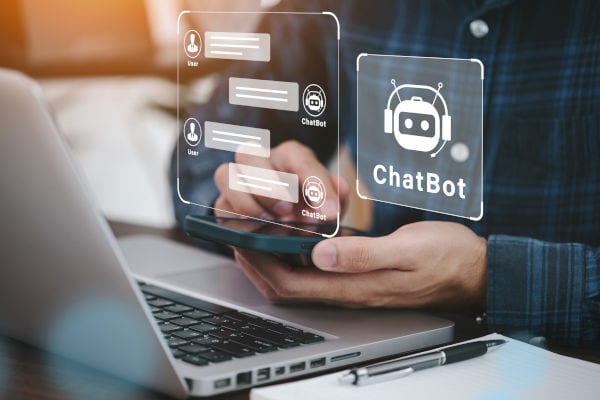 Person nutzt ein Smartphone neben einem Laptop; darüber sind grafische Chatbot-Symbole und Chatverläufe eingeblendet, die eine digitale Unterhaltung mit einem KI-Chatbot darstellen.