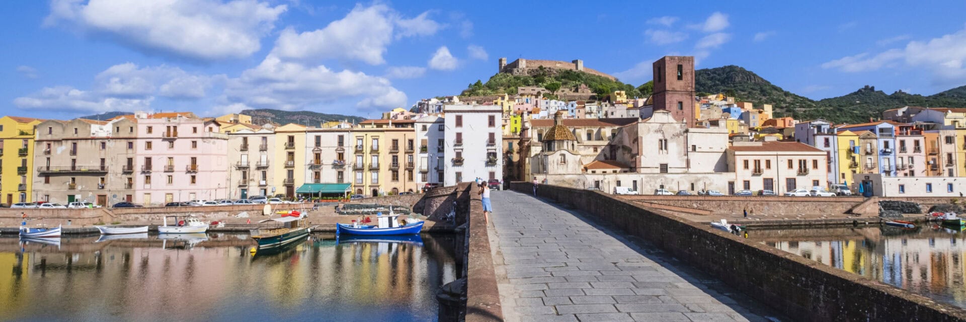 Blick über einen ruhigen Hafen mit kleinen Booten auf eine bunte Altstadt; im Vordergrund führt eine steinerne Brücke über das Wasser, im Hintergrund liegen farbige Häuser und eine Burg auf einem Hügel unter blauem Himmel.