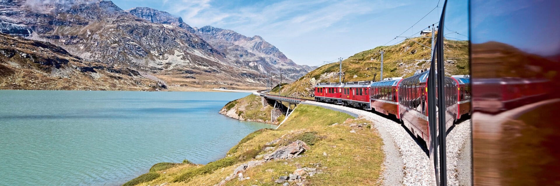 Roter Panoramazug fährt auf einer kurvigen Bahnstrecke entlang eines türkisfarbenen Bergsees, umgeben von alpinen Bergen unter blauem Himmel.